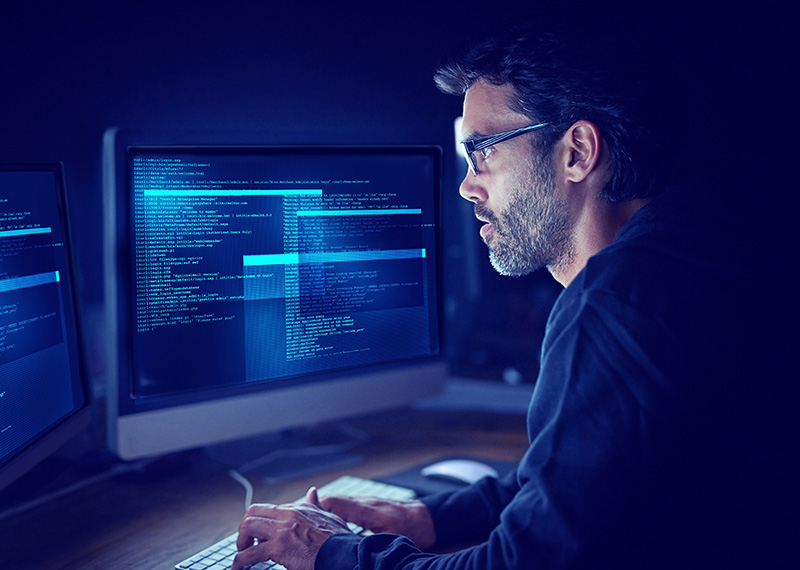 a software developer in darkly lit room viewing source code on two monitors